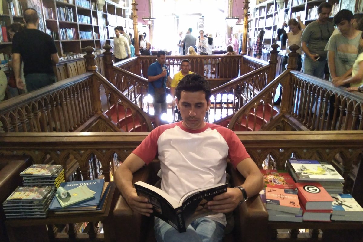 Diego Fresno sitting reading on the upper floor of Livraria Lello, with carved wooden shelves behind him